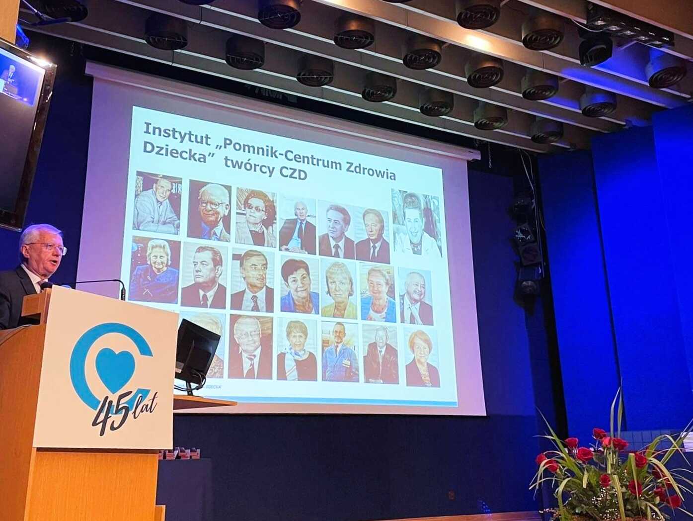 Zdjęcie sceny z dyrektorem CZD dr. n. med. Markiem Migdałem przy mównicy. W oknie sceny ekran ze zdjęciami osób związanych z CZD